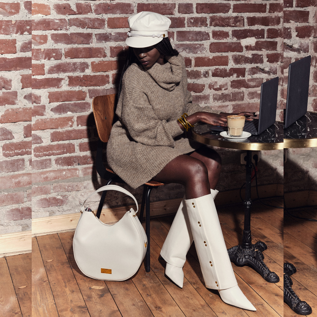 Model im Café am Laptop mit Ensemble Yve aus Handtasche, Bakerboycap und Highschaft Boots in creme 